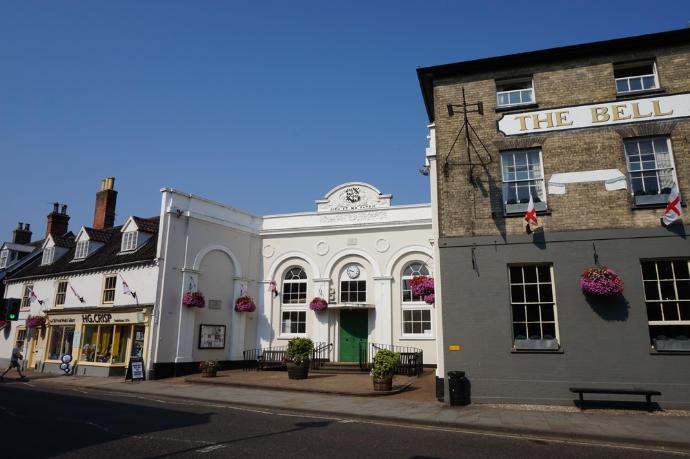A photo of a historic white public building painted white, next to a bigger Victorian building with part of the wall painted grey. Letters on the front of the building day Bell Inn. The sky is blue with no clouds. Geraniums in hanging basket hang on the front of both buildings.