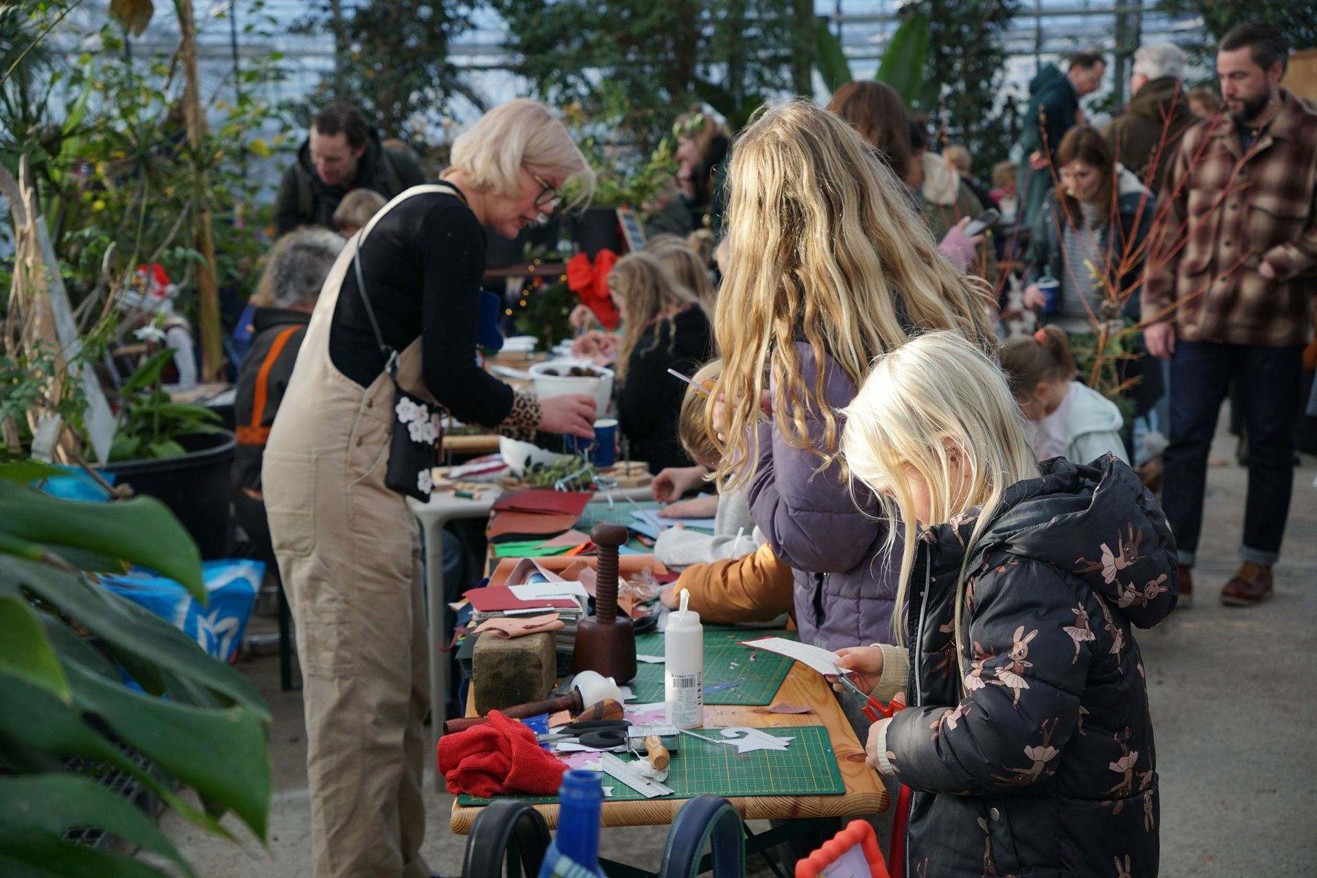 A woman with grey hair wears dungarees, and standing behind tables set up with gardening items on the tops. Children and a couple of adults looking at the items. The setting looks like a large greenhouse.